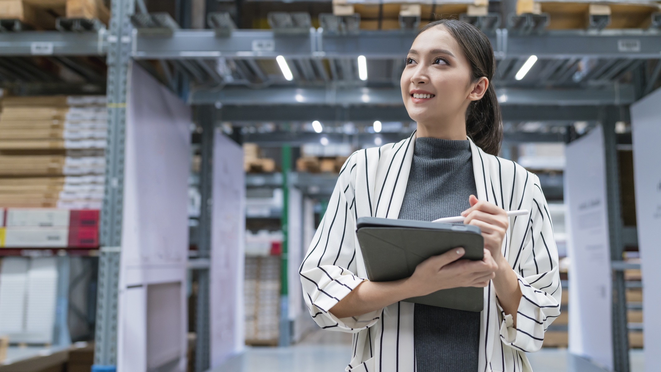 Asian woman walking through warehouse Woman smiling and checking inventory on tablet